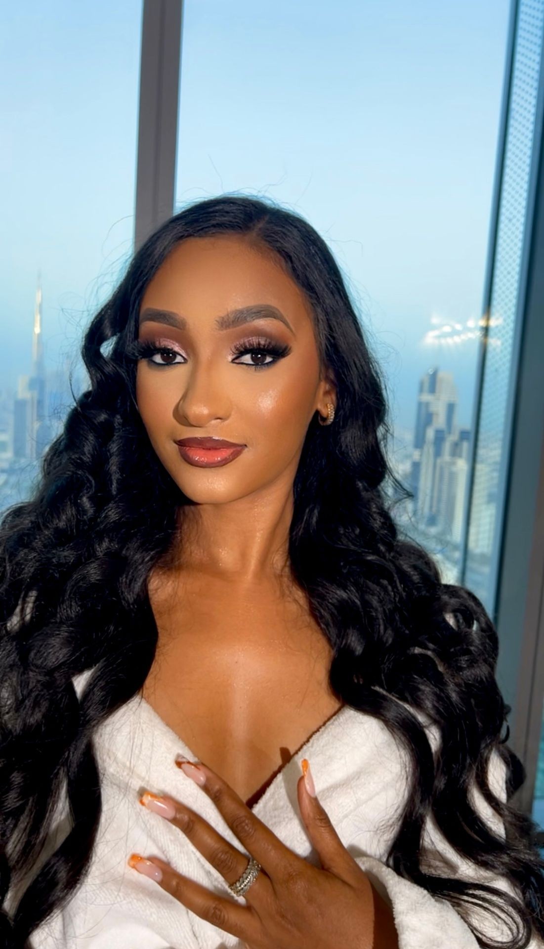Woman with long wavy hair and elegant makeup posing in front of a cityscape backdrop.