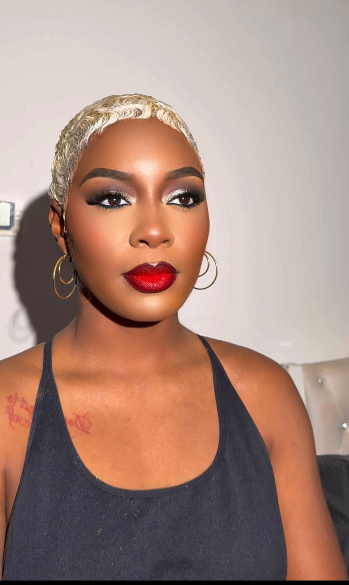 Person with short platinum hair, bold red lipstick, and gold hoop earrings wearing a black top.