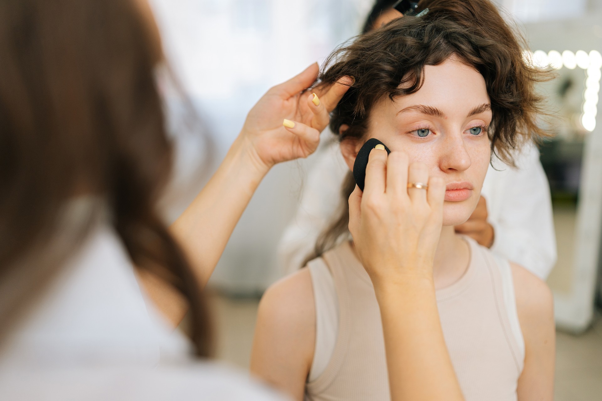 Close-up cropped shot of unrecognizable female beauty specialist using blending sponge applying foundation near woman eye.